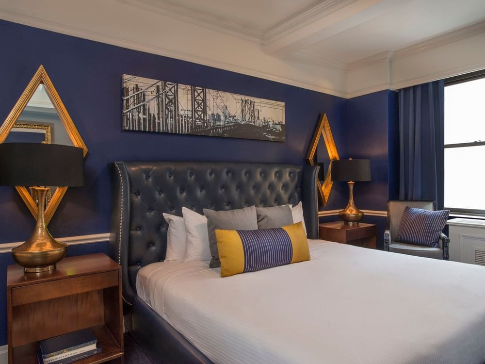 Bold blue bedroom with a leather tufted headboard & gold mirrors, and city bridge art in Randolph Suite at Warwick New York
