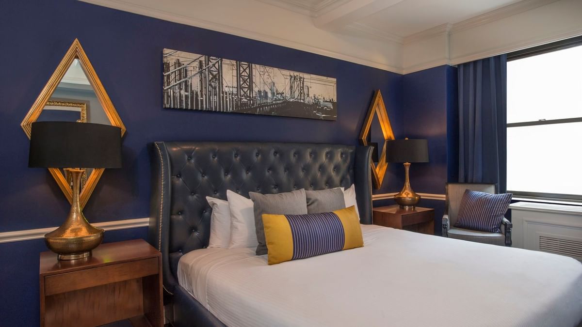 Bold blue bedroom with a leather tufted headboard & gold mirrors, and city bridge art in Randolph Suite at Warwick New York