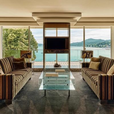 Elegant living room with two striped couches, glass table, and large windows overlooking a lake.