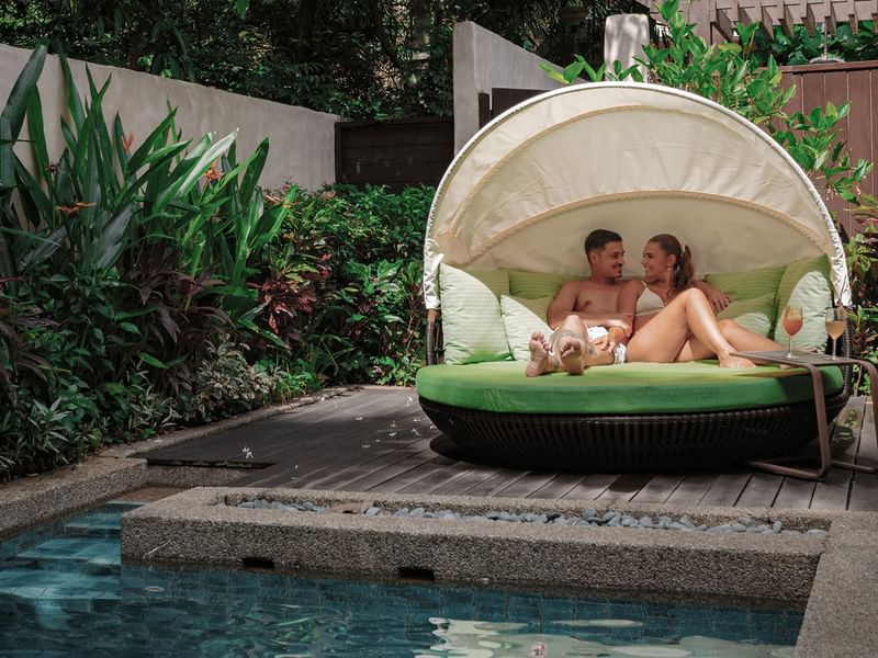 Couple relaxing on a daybed with canopy and wine near a pool, surrounded by lush plants promoting 3D2N celebrate love festive package