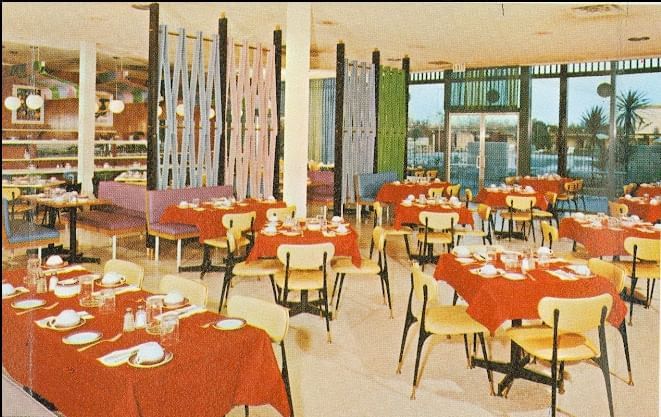 A dining area with multiple tables set with red tablecloths and yellow chairs, large windows in the background.
