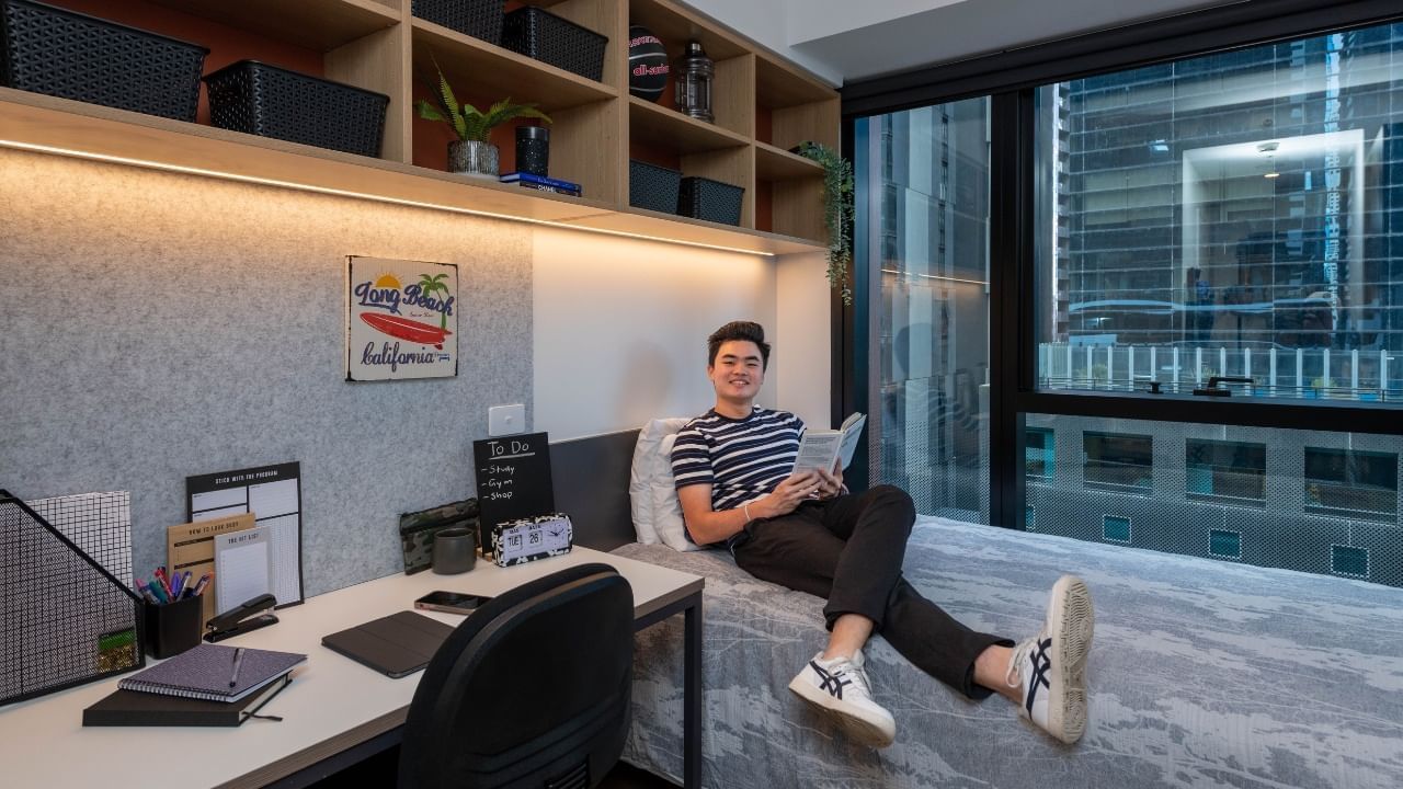 Resident in striped shirt reading a book sitting on a bed next to a desk.