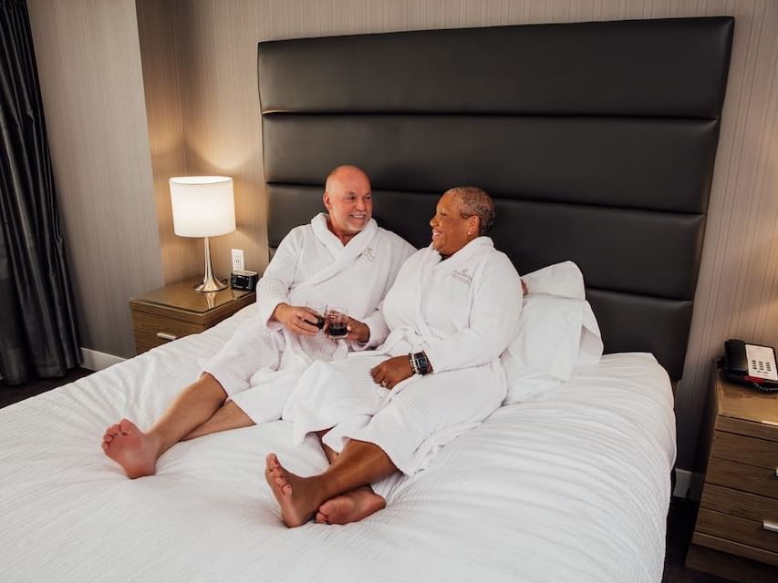 Two people in robes sitting on a bed holding wine glasses at Sandman Hotel & Suites Winnipeg Airport