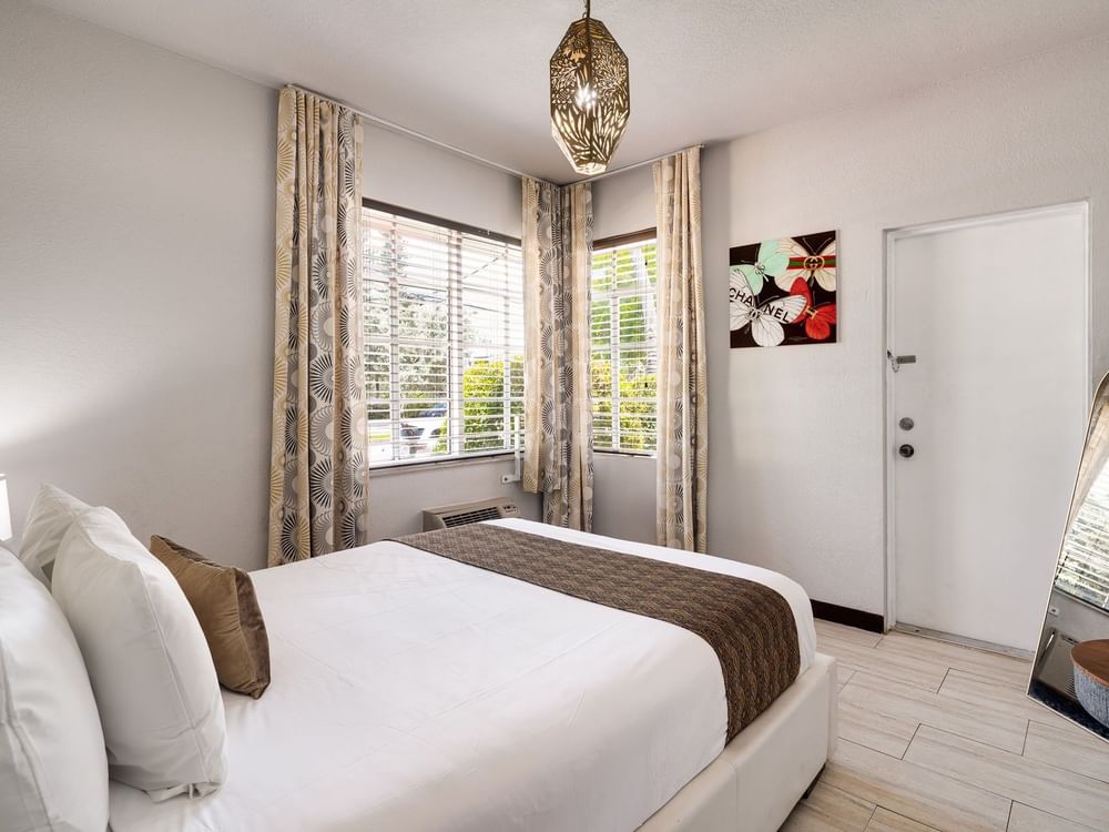 A wide-angle view of a hotel room with a bed, two windows, a door, and artwork on the wall.