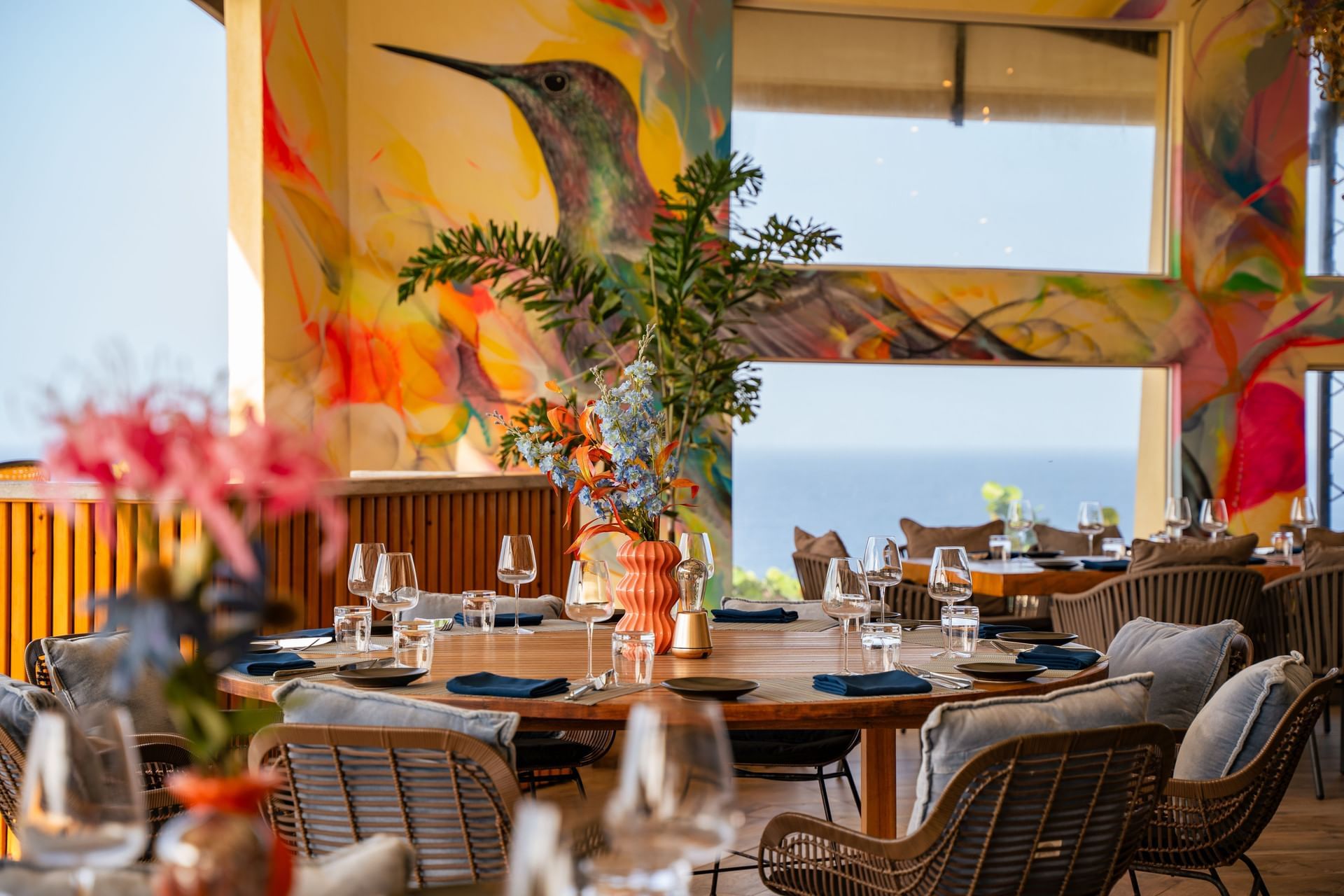 Dining area with a vibrant mural, elegant table settings, and ocean views in the Bobbie's Beach Club  at Golden Rock Resort