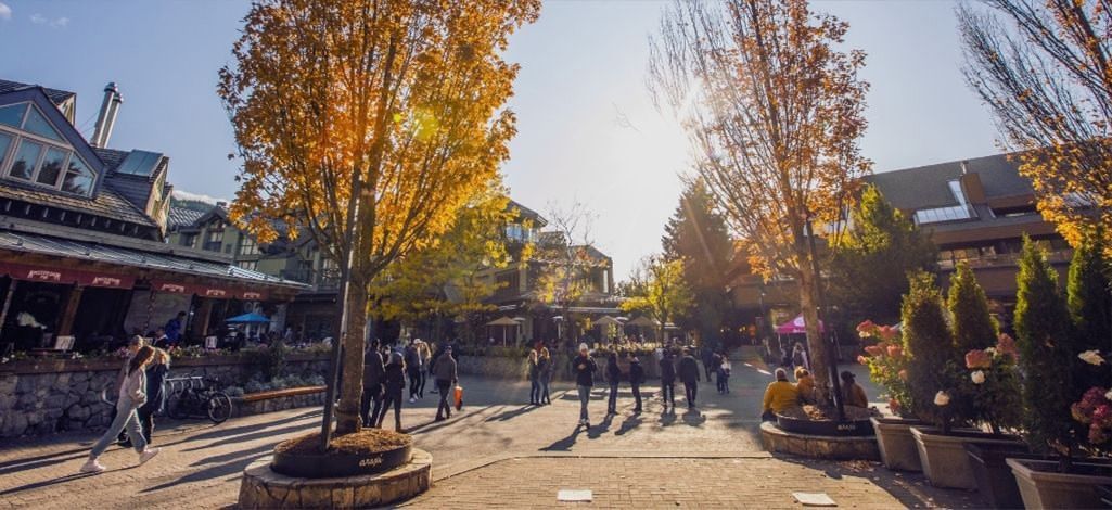 People walk through Whistler Village on a sunny autumn day, with golden-leaved trees, mountain-style buildings, and outdoor patios creating a lively atmosphere.