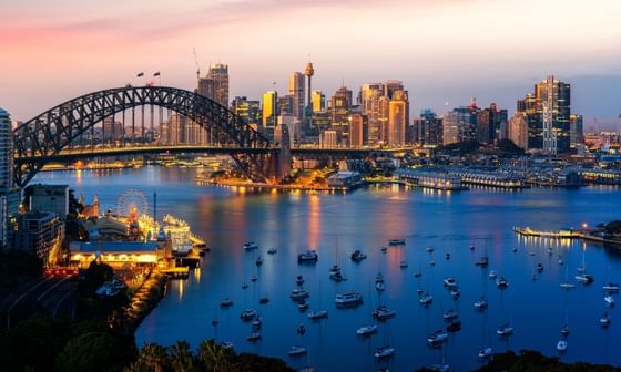 Sydney skyline at dusk, showcasing the iconic arch bridge and harbor filled with boats.