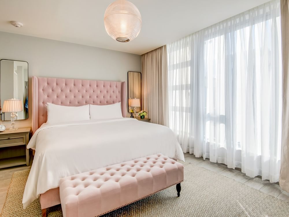 A wide view of a hotel room with a pink tufted headboard and bench, white bedding, and a large window with sheer curtains.