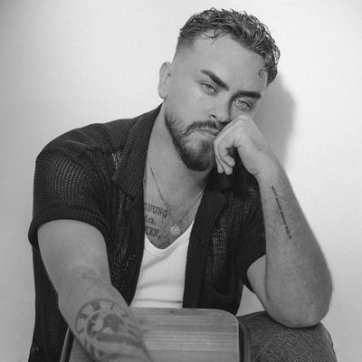 Black and white photo of a man with tattoos and a beard, sitting with a guitar.