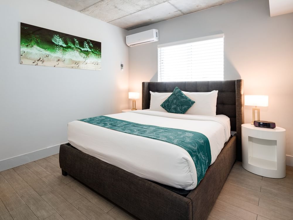 A neatly made king-sized bed with a dark upholstered headboard, white bedding, and teal accents, in a light-colored room with a window and artwork.