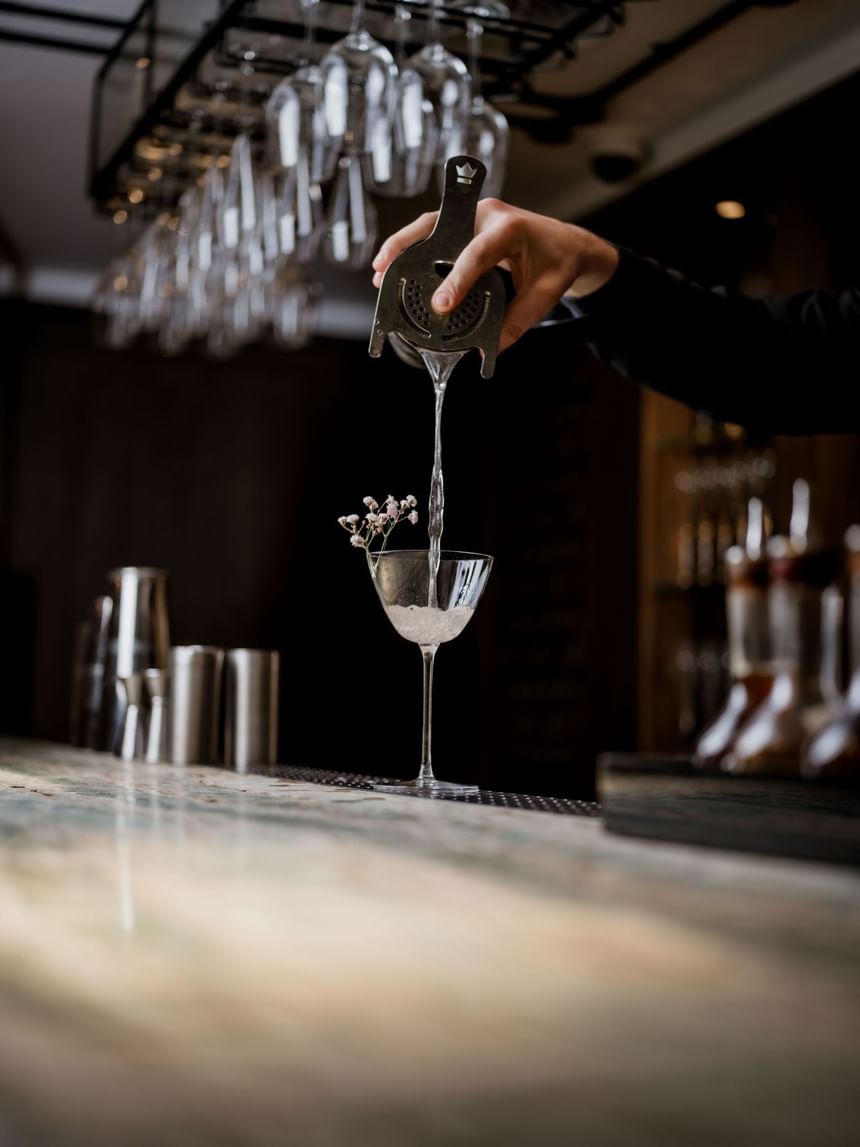 Bartender pouring cocktails into a glass and glassware hanging above, and a bar surface at The Kitchens