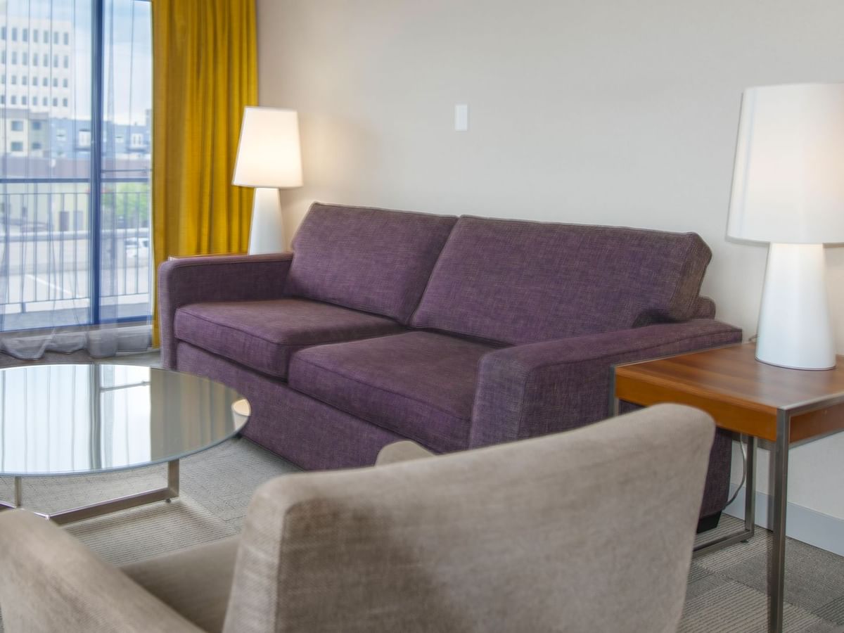 Purple couch, gray chair, glass table, and two lamps in a room with a city view.