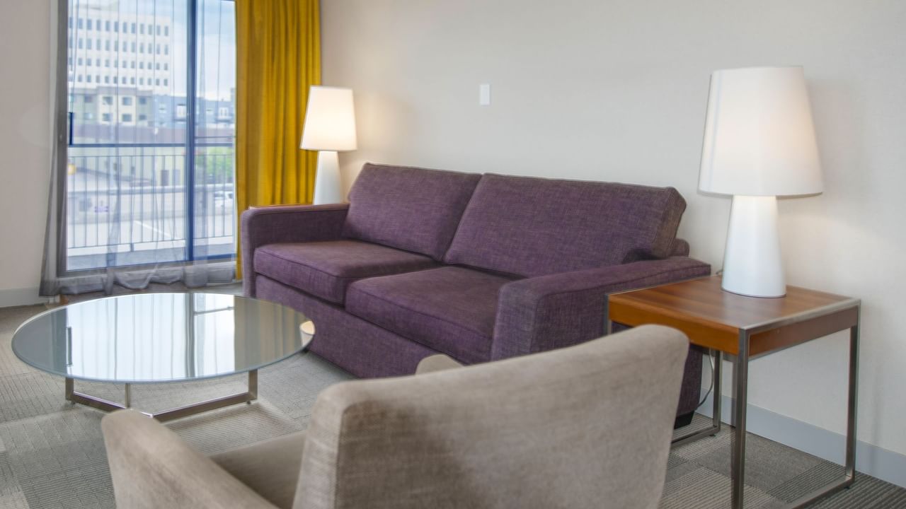 Purple couch, gray chair, glass table, and two lamps in a room with a city view.