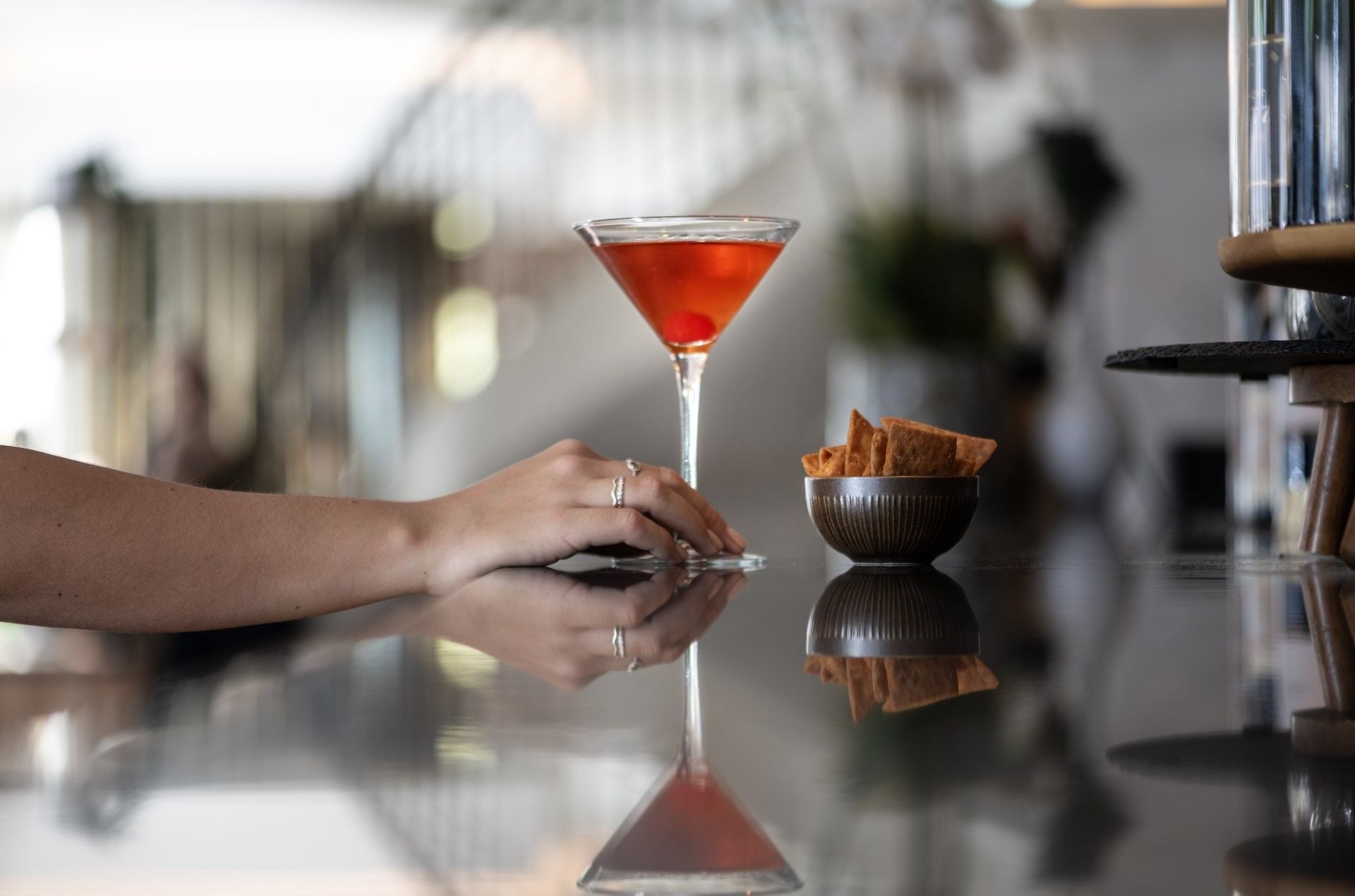 Hands holding a cherry martini on a reflective counter, by a bowl of chips in Téséo Bar at Warwick Geneva