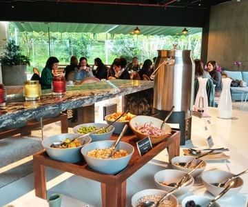 Breakfast buffet bar with condiment bowls and beverage dispenser near diners in a bright restaurant at Unika Hotels