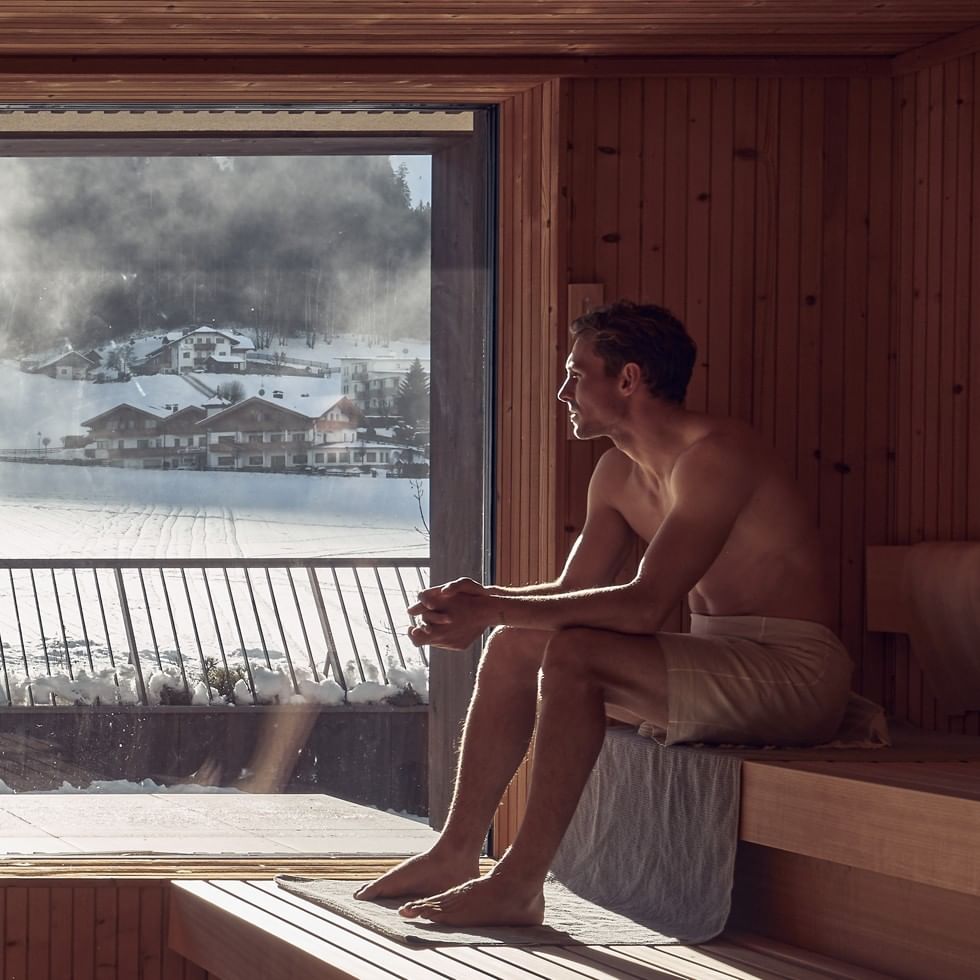 Man relaxes in a sauna with a snowy mountain view through the window.