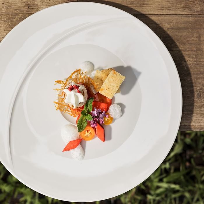 White plate with a dish containing herbs, flowers, fruits, and chips, set on a wooden surface.