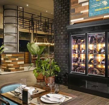 Beef Grill Club, Titanic Deluxe Lara’da bitkili, vitrin dolaplı, masalı ve sandalyeli restoran.