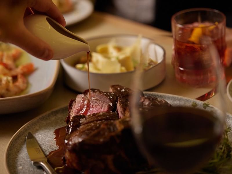 Hand pouring sauce over sliced steak with sides & a drink in Modo Mio at Crown Promenade Perth