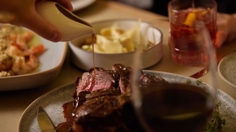 Hand pouring sauce over sliced steak with sides & a drink in Modo Mio at Crown Promenade Perth