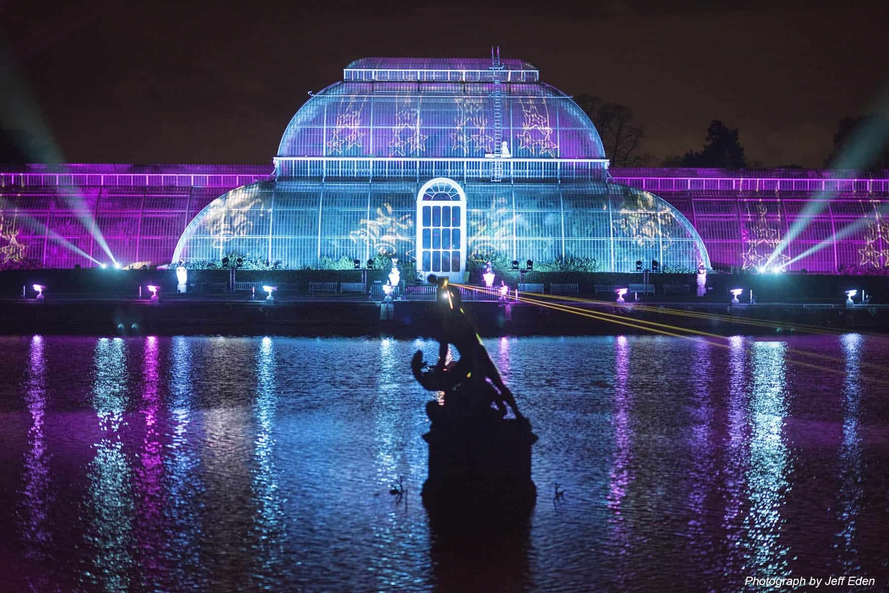 Glass conservatory illuminated with blue and purple lights reflected in pond with silhouetted figure at Kew Gardens
