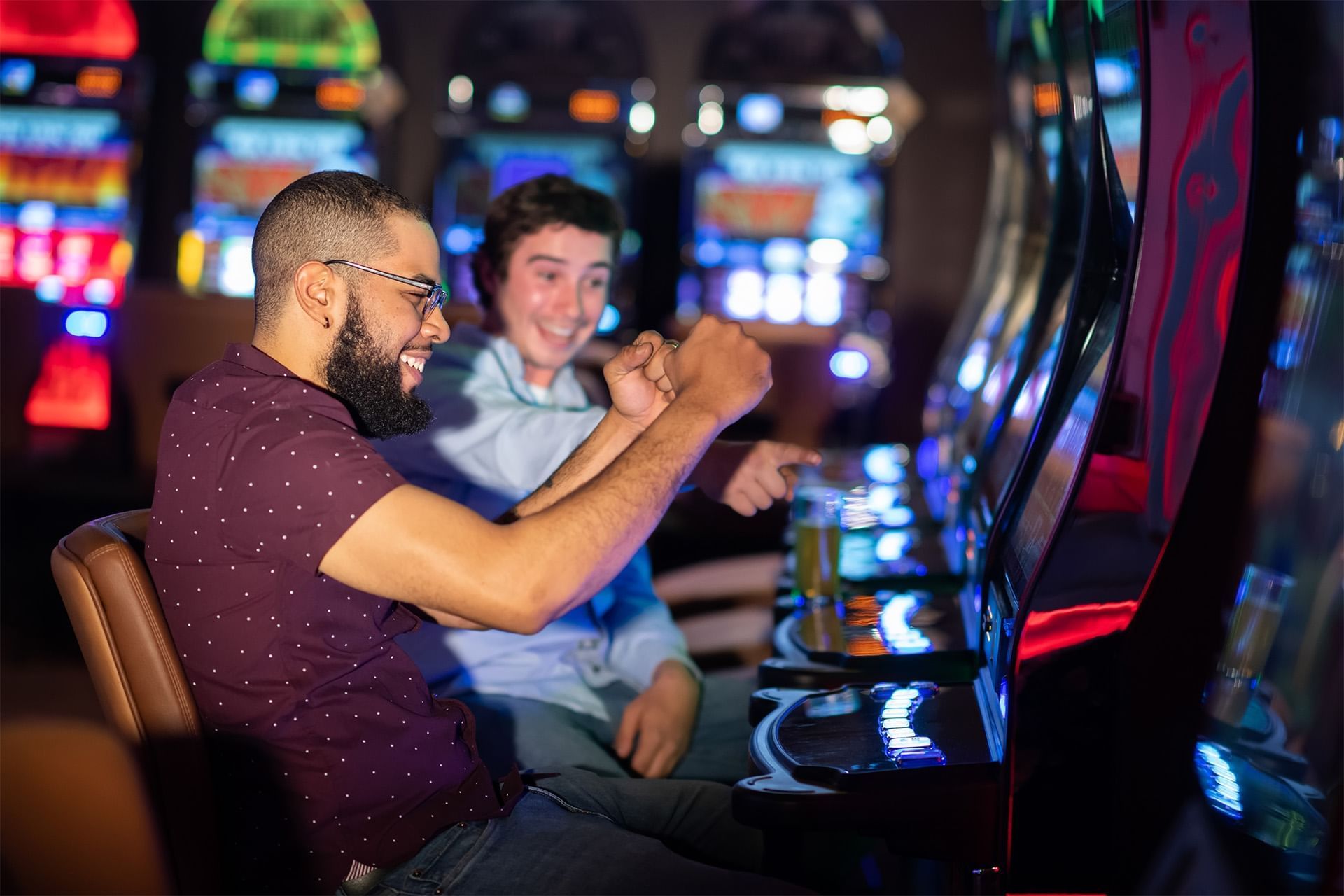 Two men celebrating excitedly beside a glowing slot machine, fists raised and pointing under casino lights at The Brook