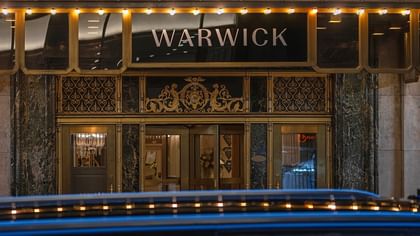 Entrance to Warwick New York hotel with car passing in front with marquee lights above, surrounding ornate gold designs