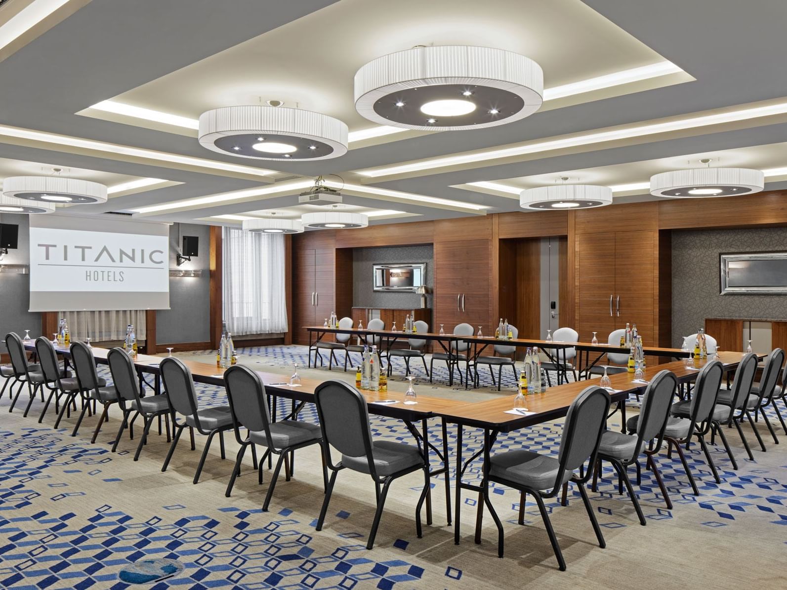 Konferenzraum mit langen Tischen, Stühlen, Projektionsleinwand und Beschilderung – Titanic Chaussee Berlin.