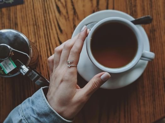 An overhead shot of a hand from a denim jacket gently touching the rim of a white mug filled with tea. A teapot, spoon, and saucer are also on the table.