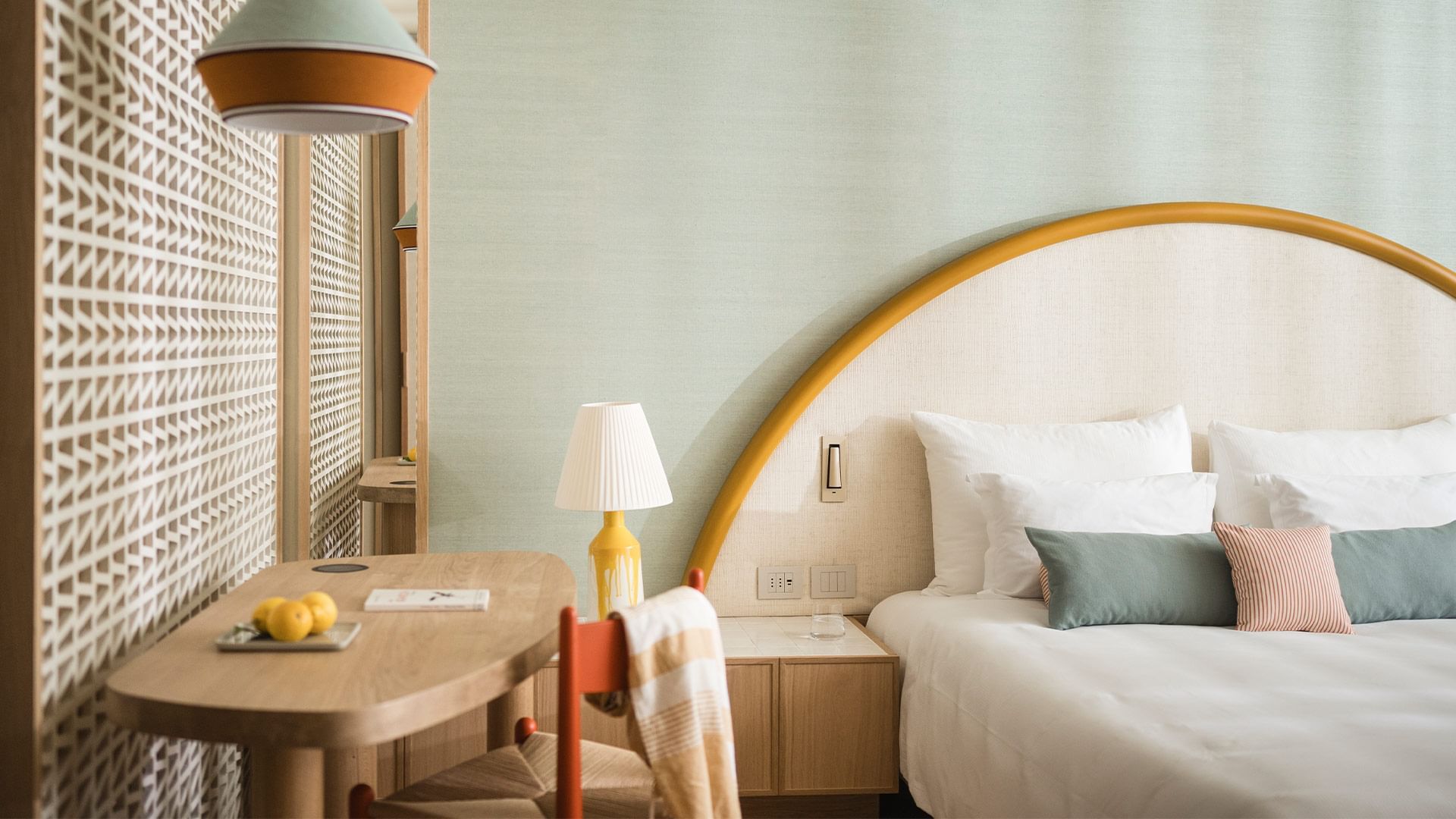 A hotel room with a bed, pillows, desk, chair, lamp, towel, and fruit on the desk.