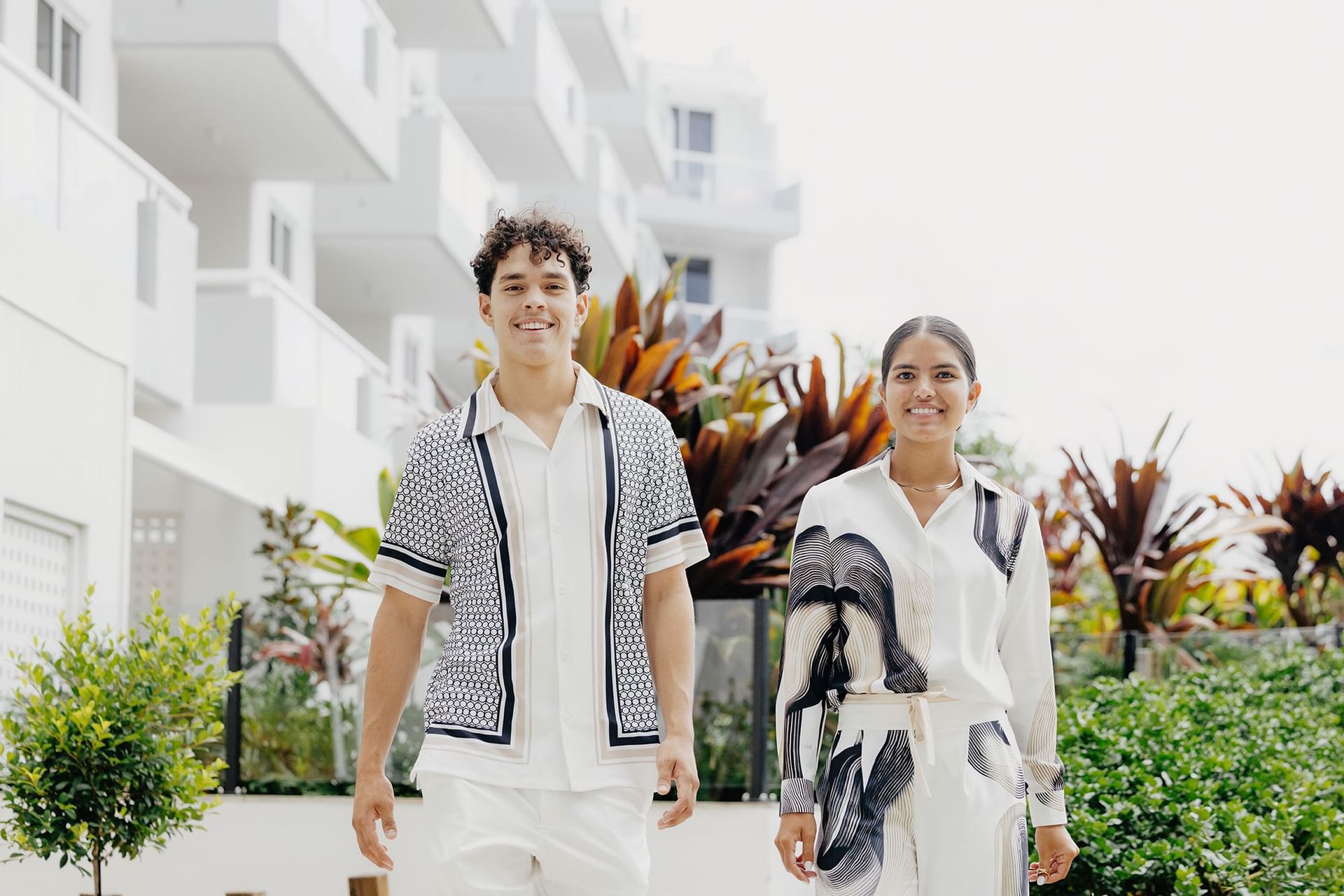 Two guests smiling and walking past lush plants and modern building at Elysium Hotels