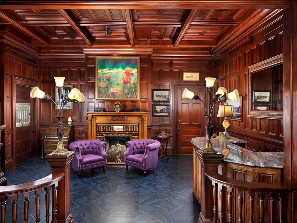 A richly decorated lobby with wooden paneling, two purple chairs, a fireplace, and a reception desk.