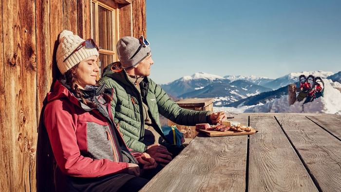 Paar genießt eine Mahlzeit auf einer Holzbank mit Bergausblick bei Falkensteiner Group