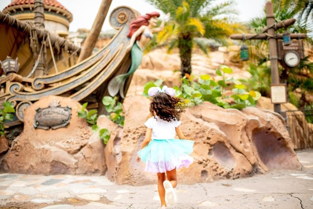 A toddler at Walt Disney World in a blue and purple tutu runs toward a Little Mermaid display.