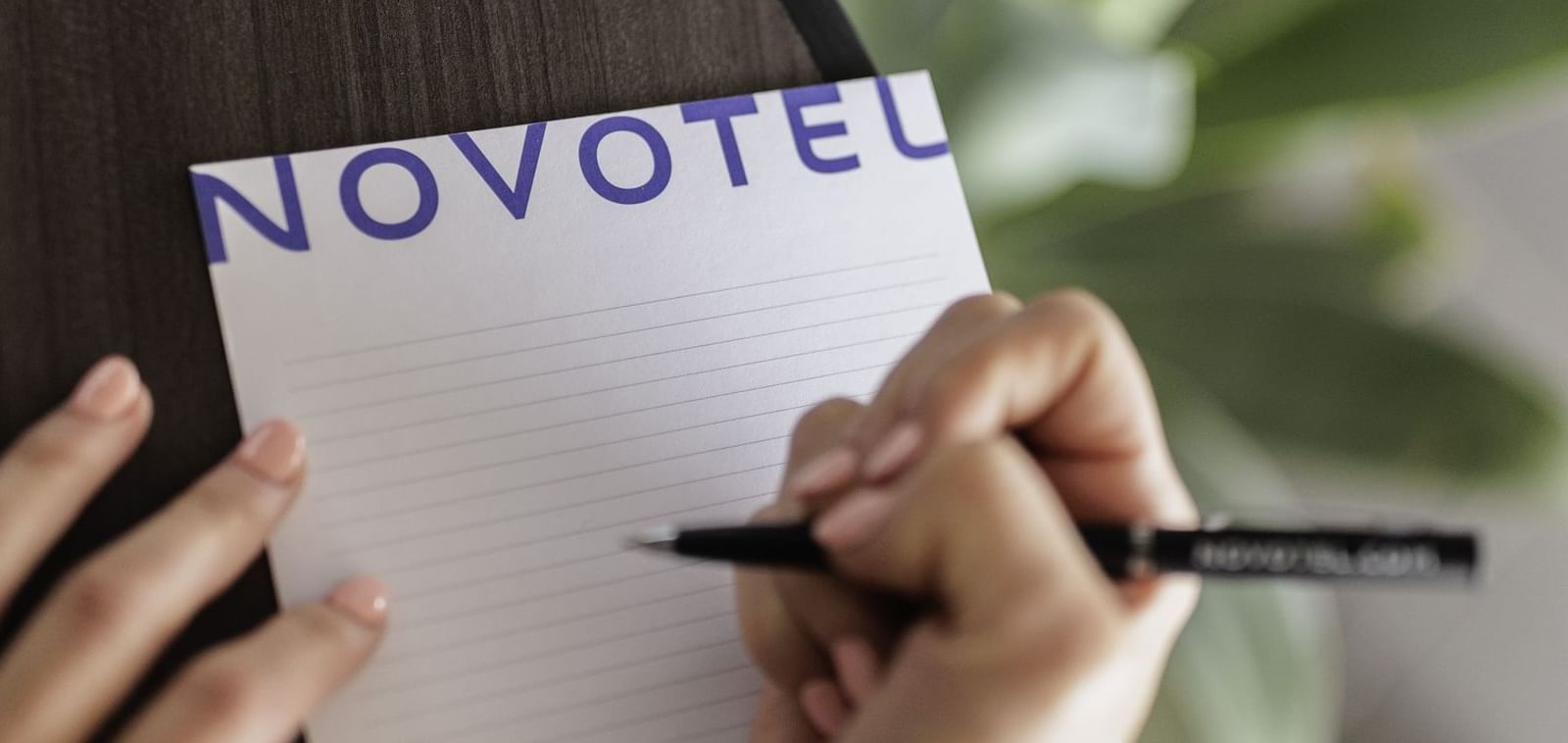 Hand holding a pen writing on a notepad with NOVOTEL printed in top set against a wooden table at Novotel Olympic Park