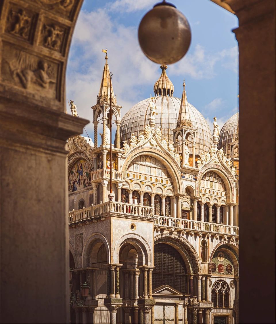 Architectural arch view of ornate Saint Mark Basilica façade and stone domes under clear blue sky near Meraviglia