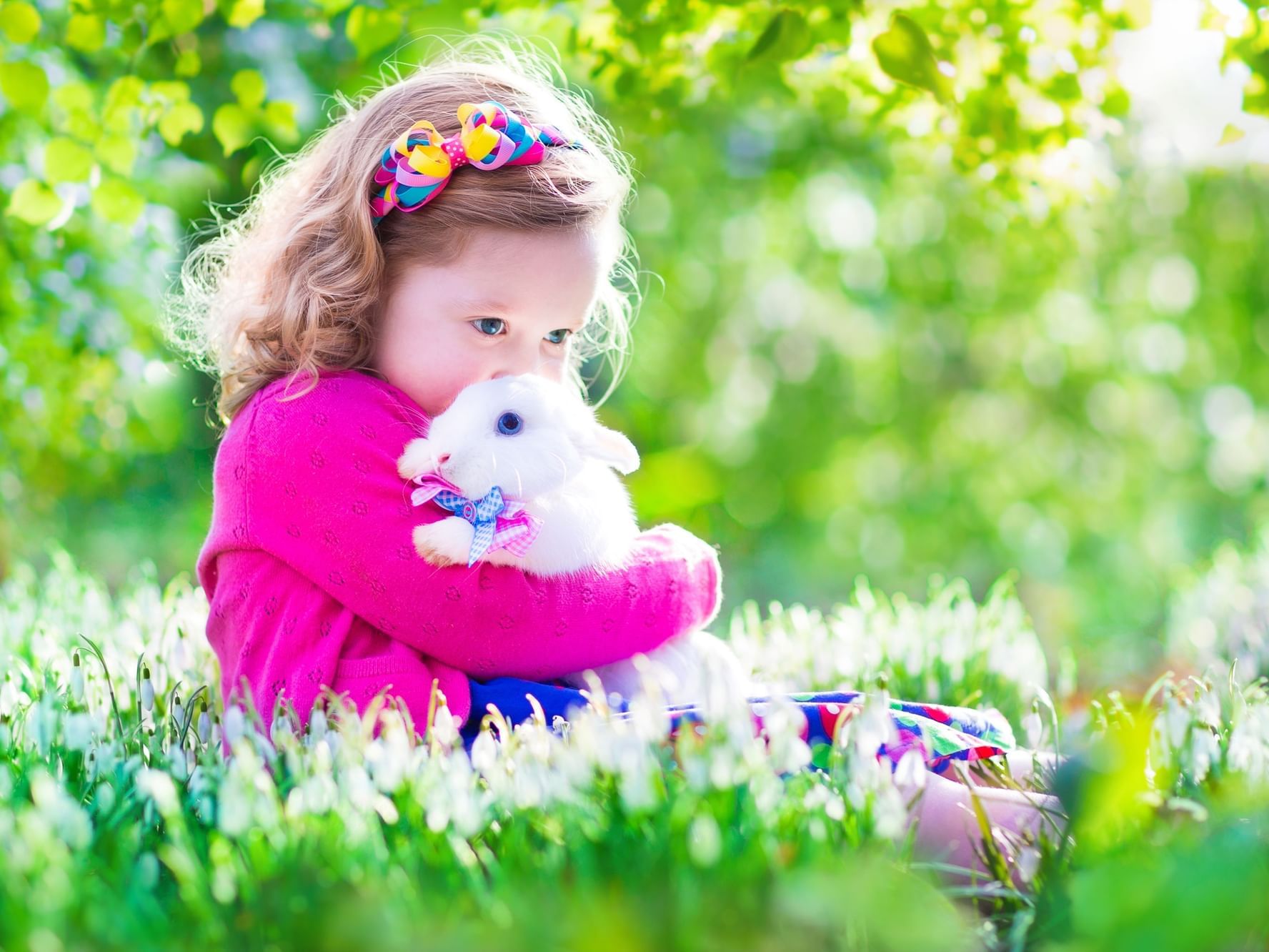 Young child sits in a grassy area at Ana Hotels, holding a white rabbit, surrounded by vibrant greenery and soft sunlight