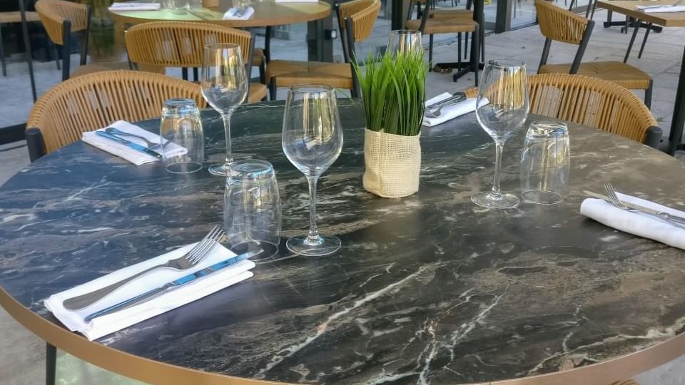 Table ronde en marbre avec chaises en osier et ustensiles de table dans un restaurant avec vue sur l'extérieur.