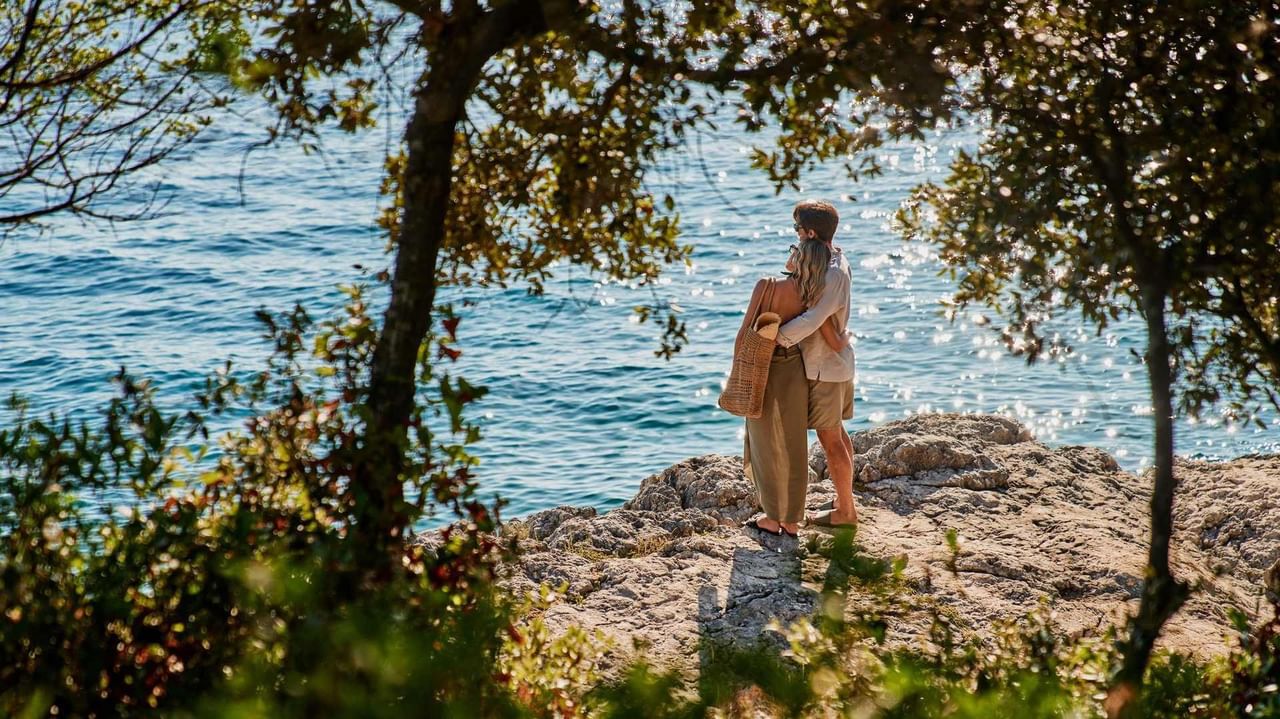 Zwei Menschen umarmen sich auf felsigem Ufer nahe einem See, umgeben von Bäumen.