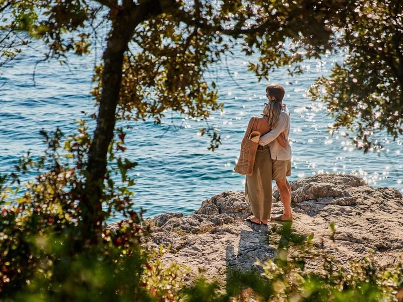 Zwei Menschen umarmen sich auf felsigem Ufer nahe einem See, umgeben von Bäumen.