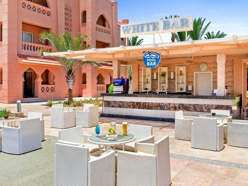 Außenbereich eines Hotels mit weißer Poolbar und Sitzbereich, umgeben von Palmen und Gebäuden.