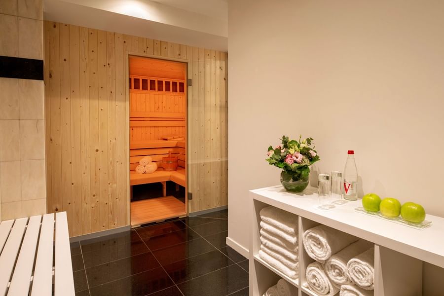 Wooden sauna with glowing interior beside white shelving unit with rolled towels, fresh flowers, and green apples.