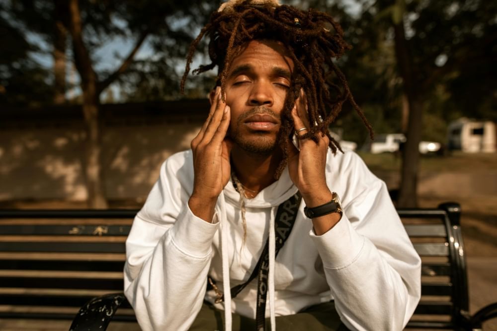 A man with his eyes closed sits on a bench holding his temples in his hands. 