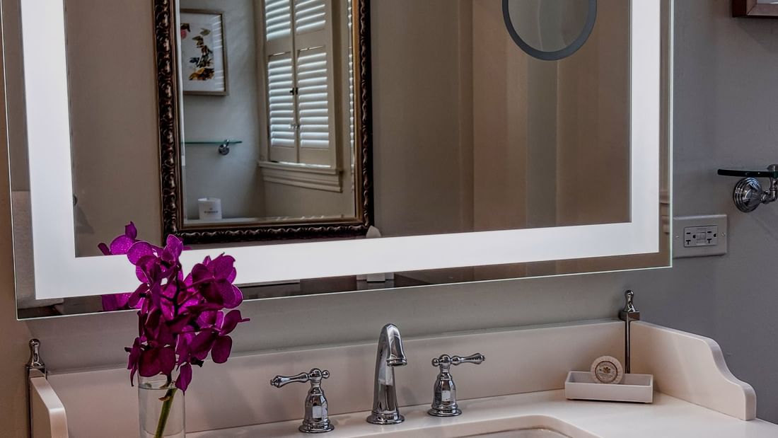 Purple orchids in a vase by a white sink under a lighted vanity mirror in Deluxe Suite at Warwick Melrose Dallas