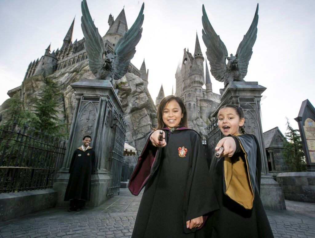 Two kids in Hogwarts robes standing in front of the gates to Hogwarts, pointing their wands at the camera.