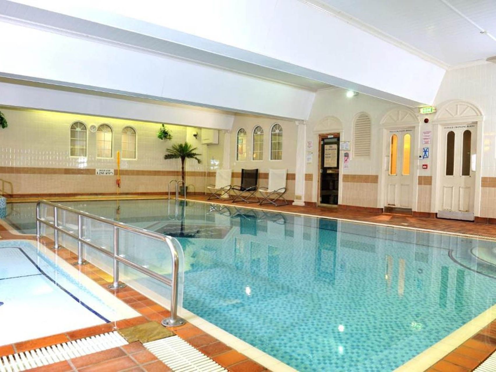 Indoor pool area with a large pool, smaller pool, and lounge chairs near a palm tree.