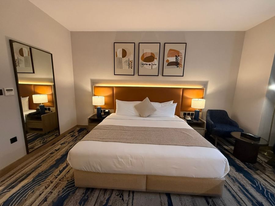 Plush bed by a blue armchair under abstract artwork in the Guest Room at Warwick Jeddah Hotel & Spa