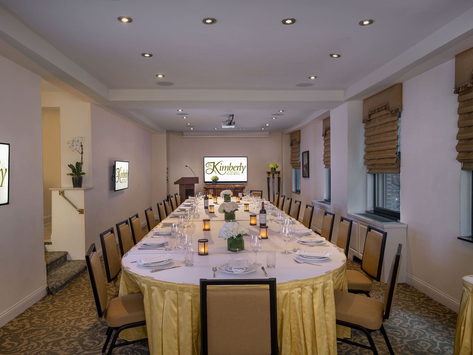 Long oval dining table set with orchids between chairs facing the TV screen in The Conference Room at The Kimberly Hotel