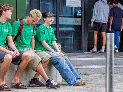 Three young people in matching green shirts sitting on a concrete ledge outside a building entrance with glass doors.