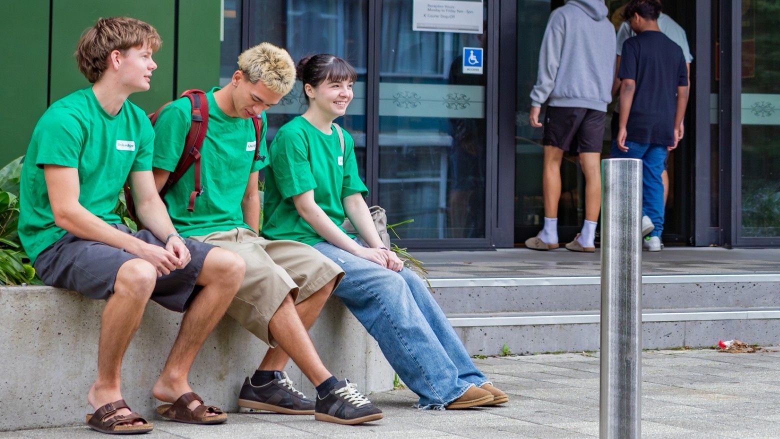 Three young people in matching green shirts sitting on a concrete ledge outside a building entrance with glass doors.